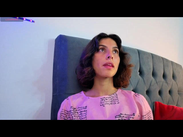 LuisaNinaSky, avec ses cheveux bruns bouclés, portant une chemise rose avec une écriture noire, assise contre une tête de lit bleue capitonnée, regardant vers le haut avec une expression légèrement confuse..