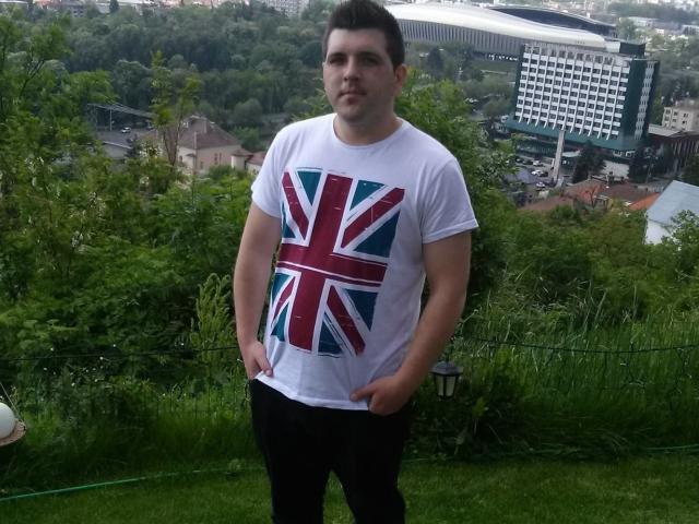 JasonJacks, chubby with short dark hair, wearing a white t-shirt with a Union Jack, hands in black pants, standing on a grassy hill, green trees in background.
