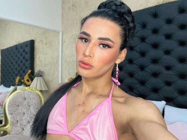 Nohemi, mit rosa Bikini-Oberteil, geflochtenen Haaren, rosa Ohrringen und einem verführerischen Gesicht, zeigt kleine Brüste und Achselhaare und steht vor einem schwarzen, gepolsterten Kopfteil..