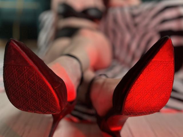 GoddessSophie69's red, diamond-patterned soles, pointed heels, and crossed legs are in the foreground, with her striped skirt and black bra slightly blurred in the background.