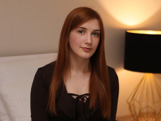 PollyFoxy has long red hair, fair skin, and small tits. She's wearing a black top with a lace-up front, sitting in a softly lit room with a black lamp.