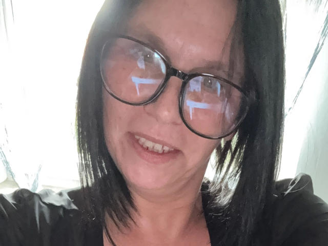 NsTempel, con capelli neri di media lunghezza, che indossa occhiali, sorride con le labbra leggermente dischiuse, riflesso di luce blu negli occhiali, indossa una camicia nera, sfondo bianco..