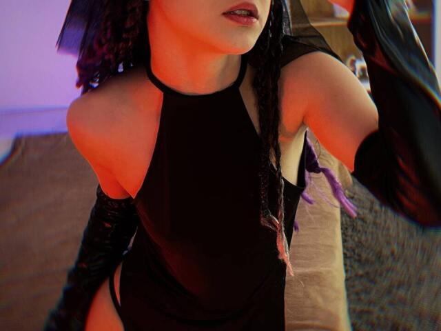 SimaWendy in a black halter top, showing bare shoulders, with purple braided hair, and black glossy arm sleeves, holding a black object, with a red-orange light.