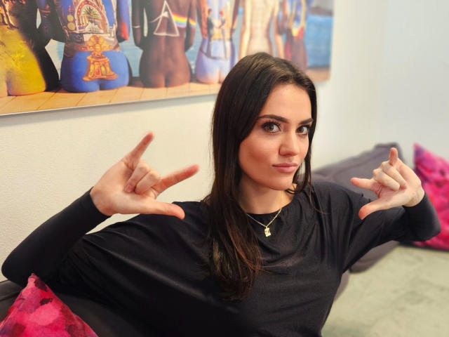 MiaLou-hot with long dark brown hair, wearing a black long-sleeve shirt, has both hands raised with fingers pointing, slightly smiling. She has a gold necklace with a small pendant. The background has a colorful painting with multiple figures, mostly in blue and yellow. Pink pillows are on a gray couch. The image is brightly lit with a focus on MiaLou-hot's expressive face and hands.