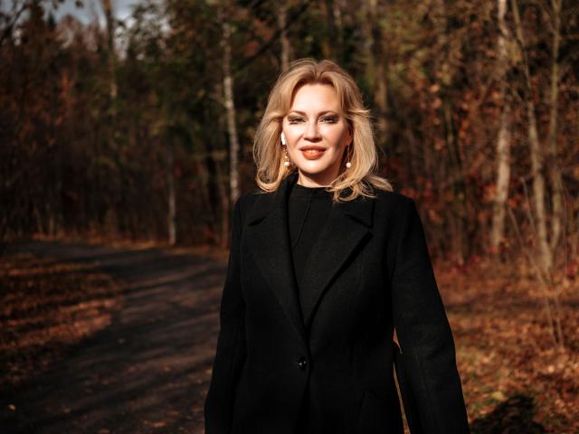 NatalyFoxy69 with blonde hair, wearing a black coat, standing on a forest path, smiling, showing off her ass in the shot, in a warm, sunlit, autumn setting.