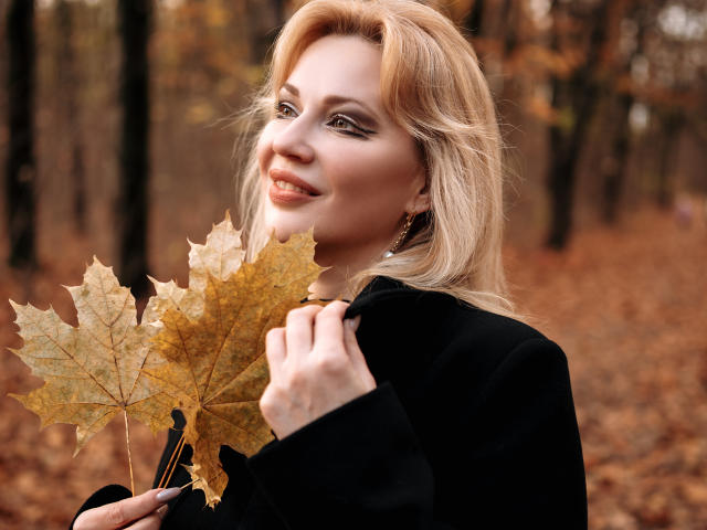 NatalyFoxy69 with blonde hair, holding autumn leaves, wearing a black coat, smiling, her tits slightly visible under the coat, in a forest with orange and brown leaves.