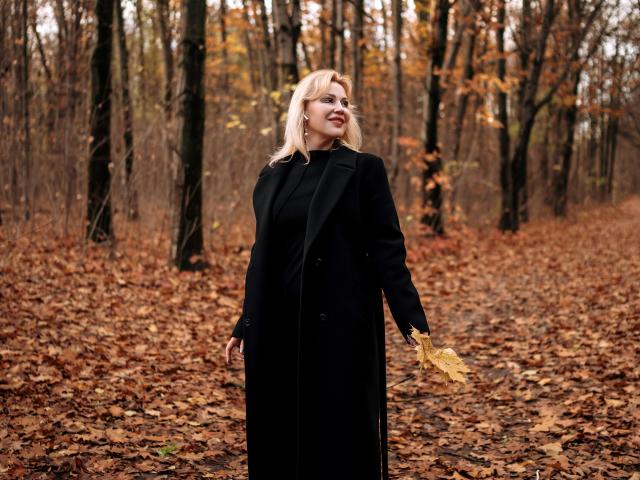NatalyFoxy69, blonde woman with medium tits, black coat, holding yellow leaf, standing on brown leaf-covered forest path, smiling, looking to the side, autumn, forest background.