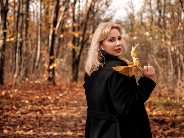 NatalyFoxy69 with blonde hair, holding yellow leaves, wearing a black coat, looking back with a slight smile, standing on a forest path covered in brown leaves.
