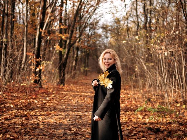 NatalyFoxy69 with blonde hair, black coat, holding yellow and white leaves, standing on a leaf-covered path in a forest, autumn colors, looking at camera, soft light, cute.