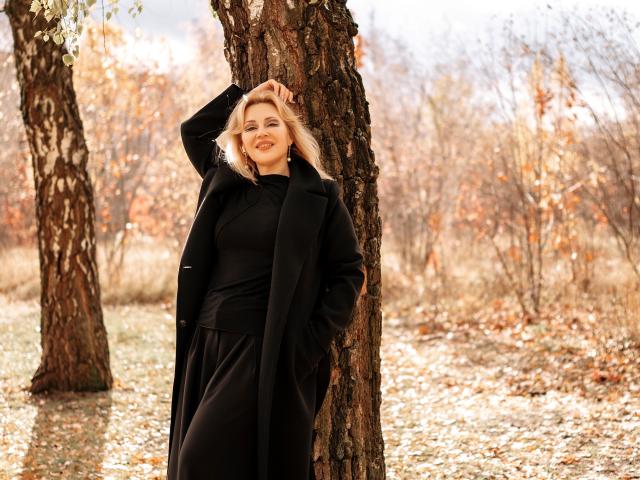 NatalyFoxy69 with blonde hair, black coat, and black dress, standing against a tree, hand in coat, smiling, autumn leaves, soft sunlight, trees in background.
