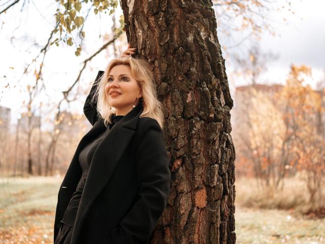 NatalyFoxy69 leans against a tree with rough bark, wearing a black coat, blonde hair, and a slight smile, hand touching her head. Autumn leaves in the background.