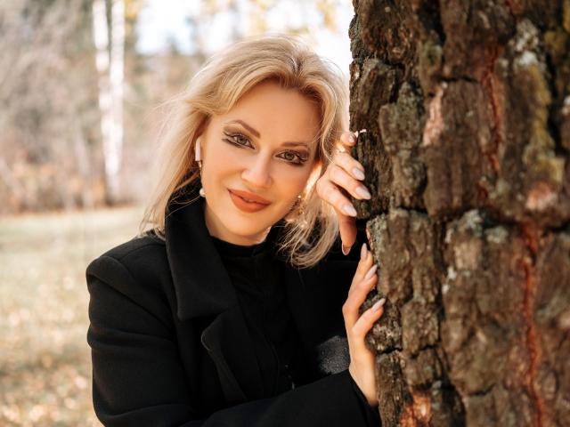 NatalyFoxy69 with blonde hair, black coat, and earrings, leaning against a tree, smiling, holding it with her right hand, showcasing her white nail polish and cute face.