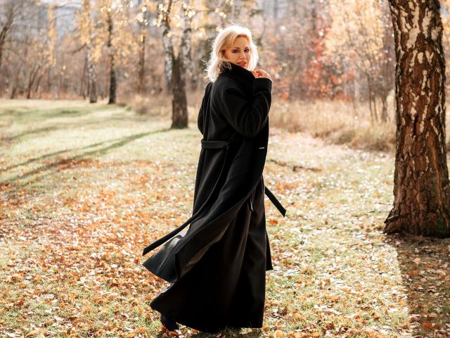 NatalyFoxy69 stands in a forest, wearing a long black coat, with blonde hair and a shy smile, holding the collar, her ass slightly visible under the coat.