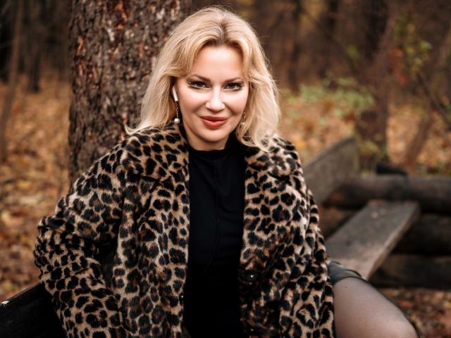NatalyFoxy69 with blonde hair, wearing a leopard fur coat, black top, and black tights, sitting on a wooden bench, smiling, with a tree in the background.