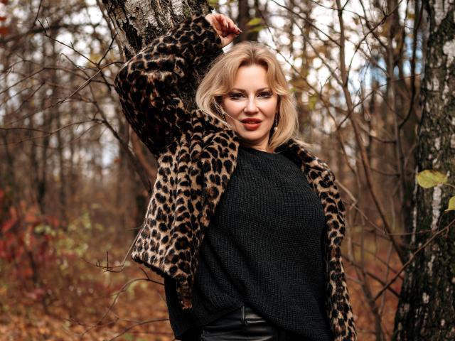 NatalyFoxy69 with blonde hair, leopard jacket, black sweater, and black leather pants, leaning against a tree in a forest, her arm raised, sexy and confident.