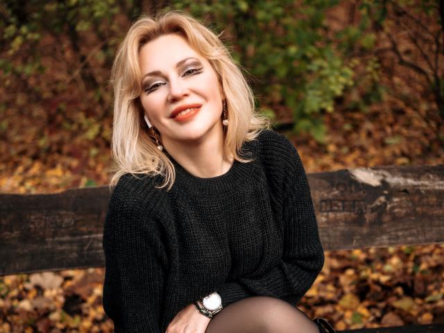 NatalyFoxy69 with blonde hair, smiling, wearing a black knit sweater, earrings, and a watch, sitting on a wooden bench, autumn leaves in the background.