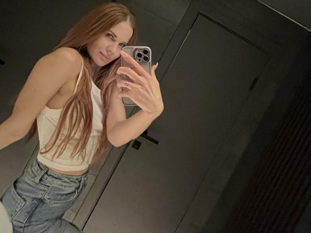 LunaLickies with long red hair, smiling, wearing a white tank top and blue jeans, taking a mirror selfie with a smartphone in a dimly lit bathroom.