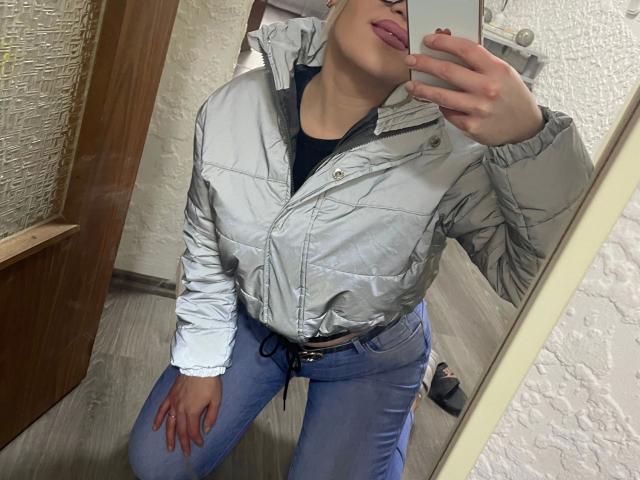 HerrinKristina92-hot in a silver jacket, black shirt, and blue jeans, taking a mirror selfie with her phone, showing her pouty lips and pink lipstick. Her hand is on her thigh.