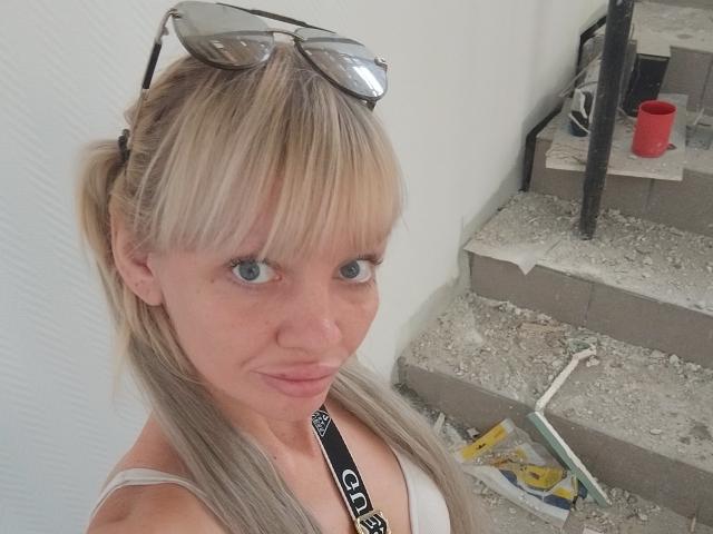 CamileRay with blonde pigtails, blue eyes, and eyeglasses on head, wearing a white tank top, standing next to a construction site with stairs and tools.