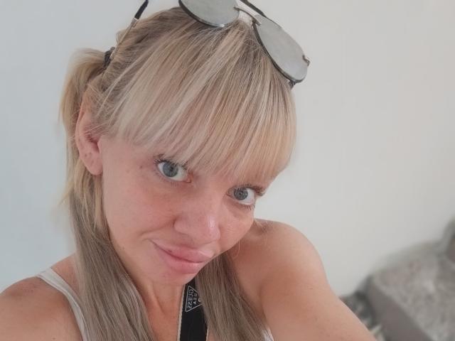 CamileRay has blonde hair in a ponytail, blue eyes, and is wearing sunglasses on her head. She's smiling slightly with a pinkish nose and wearing a white tank top.