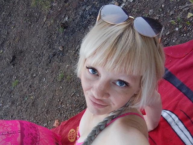 CamileRay with blonde hair, blue eyes, and pink bra, wearing sunglasses on her head, lying on a red towel, looking up with a slight smile.