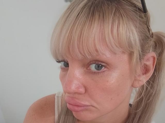 CamileRay has blonde hair in a ponytail, blue eyes, and puffy pink lips with a slightly annoyed expression. She's wearing a white tank top, with a black hairclip.