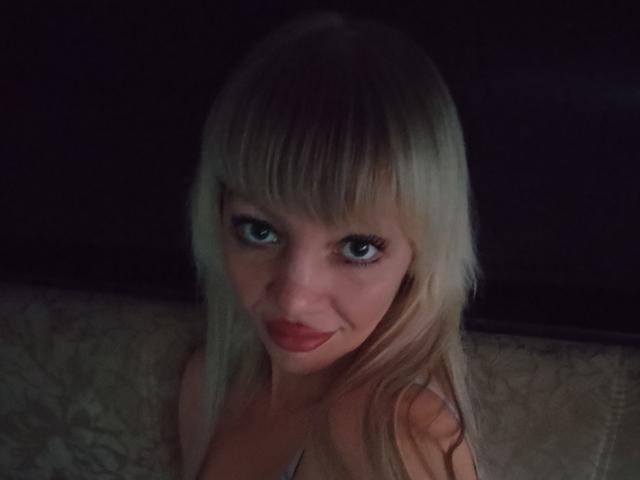 CamileRay with blonde hair, blue eyes, and red lipstick, smiling at the camera, showing her tits, in a dimly lit setting.