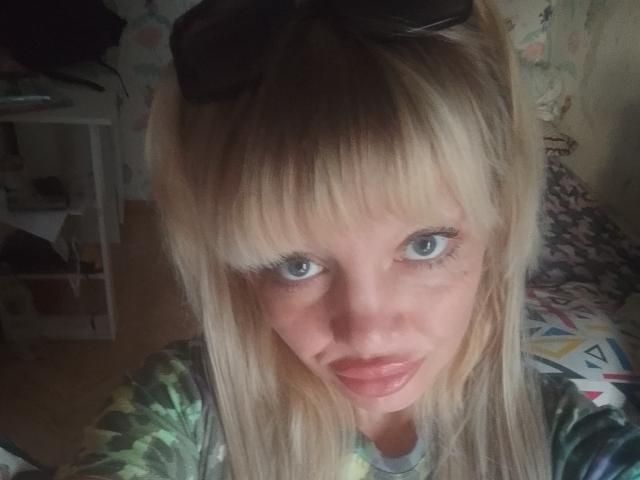 CamileRay has blonde hair, blue eyes, and pouty lips. She's wearing black sunglasses on her head and a green camouflage shirt. Her long eyelashes stand out.