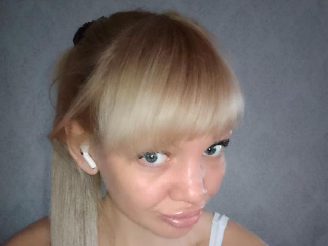 CamileRay has blonde hair with a side ponytail, blue eyes, white earbuds, and a glossy pinkish lip. Her small nose is visible. She wears a white top.