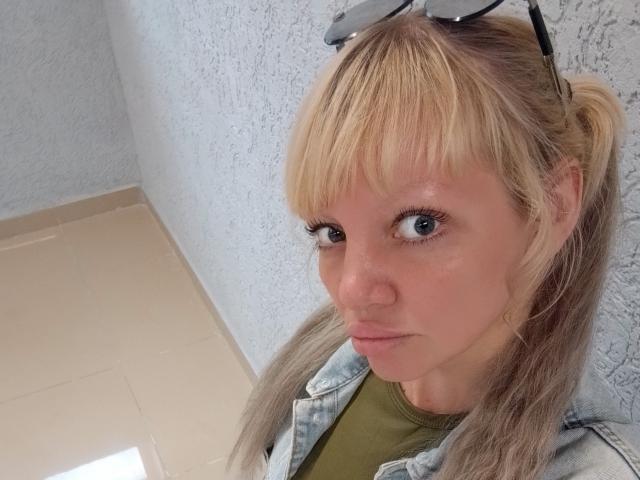 CamileRay has blonde hair in a ponytail, blue eyes, and is wearing a green shirt with a denim jacket, standing against a textured white wall. Her eyelashes are long.