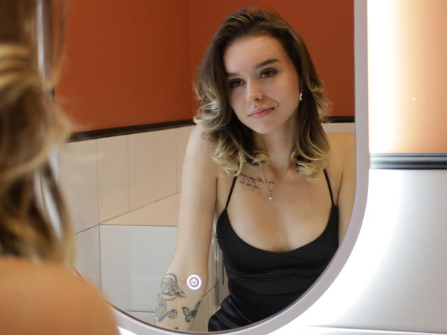 EmelyHailey with blonde, wavy hair, black dress, small tits, tattoo on right arm, smiling at mirror, white wall, orange wall, and subtle makeup. Reflection visible.