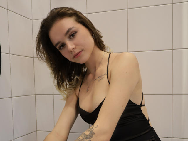 EmelyHailey with shoulder-length brown hair, wearing a black spaghetti-strap top, showing her small tits and tattoo on her right arm, sitting in a white-tiled room.