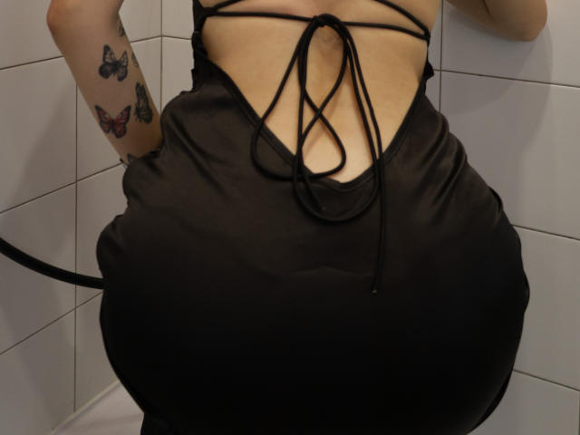 EmelyHailey's black, strappy, backless dress reveals her bare, tan-lined ass and a small, visible, cleft. Her arm, with butterfly tattoos, is partially in frame.