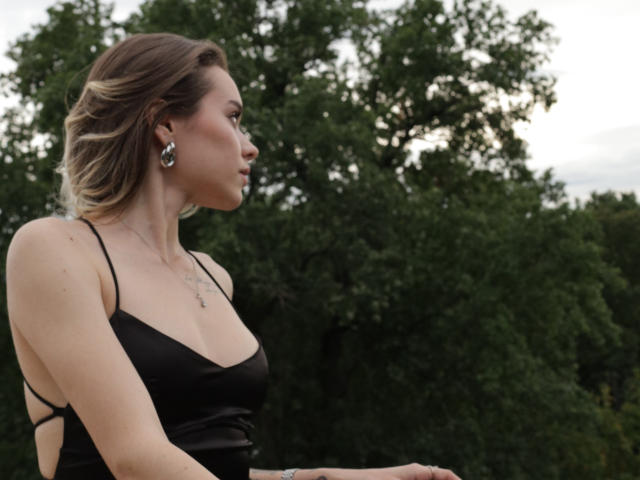 EmelyHailey has blonde hair, black dress with strappy sides, small tits, wearing silver earrings, and a necklace, looking sideways in a photo with green trees in the background.