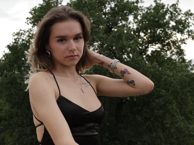 EmelyHailey with medium tits, wearing a black dress, has blonde hair, a nose ring, and tattoos on her arm, standing outdoors with green trees in the background.