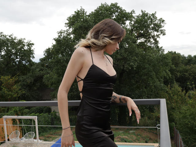 EmelyHailey with blonde ombre hair in a black dress with side cutouts, showing her small tits and tattoo on her right hand, standing outside on a balcony.