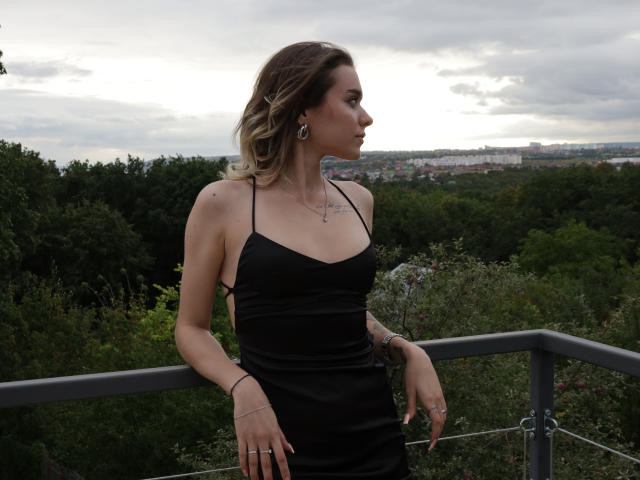 EmelyHailey in a black dress, standing with her ass slightly visible, looking to the side, blonde hair, earrings, bracelet, ring, cloudy sky, green trees, metal railing, hands down.