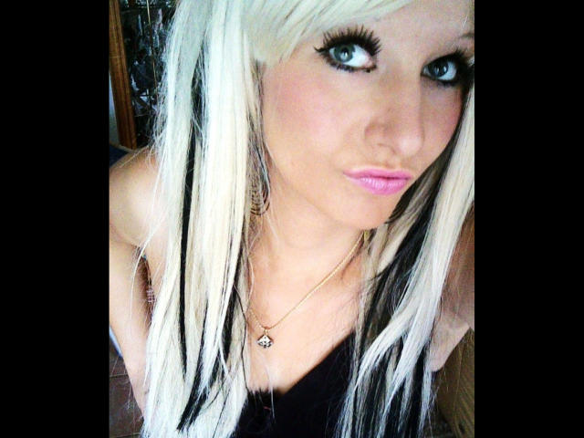 Kitty-Deluxe-hot with long, platinum blonde hair, blue eyes, pink lipstick, black eyeliner, black top, gold necklace, and a small, cute ass, looking directly at the camera.