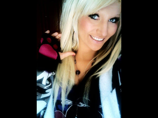 Blond Kitty-Deluxe-hot with big ass, smiling, wearing black and white plaid shirt, pink gloves, holding hair, white earrings, and necklace, in bright photo with dark background.