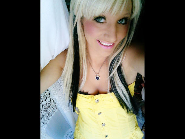 Kitty-Deluxe-hot with blonde and black hair, blue eyes, smiling, wearing a yellow button-up top, showing her tits, black bra, necklace, and lace-covered arm.