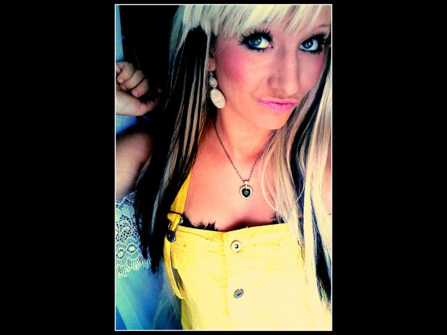 Kitty-Deluxe-hot with blonde-black-streaked hair, blue eyes, pink lipstick, yellow button-up dress, black bra, necklace, and pearl earrings, showing off her tits.