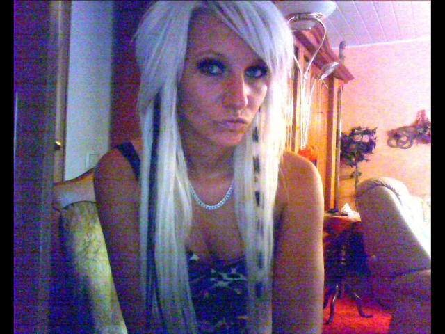 Kitty-Deluxe-hot with long white hair, blue eyes, and a pearl necklace, looking sultry, wearing a black lace top, in a dimly lit room with a green chair.