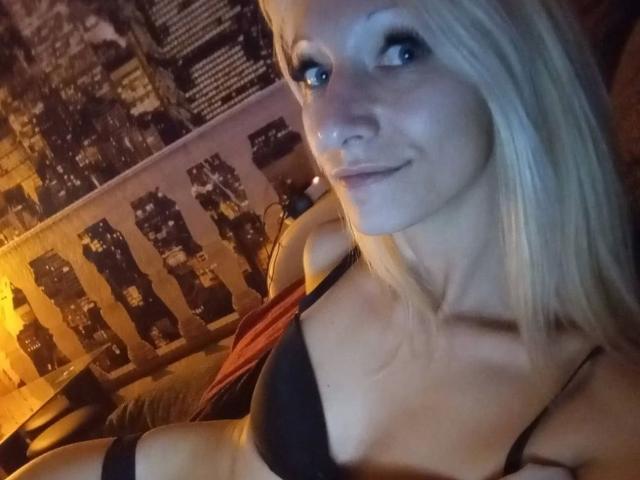 Kitty-Deluxe-hot with blonde hair, blue eyes, and black bra, showing her tits, smiling in a dimly lit room.