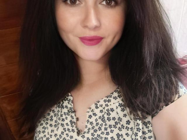 FortuneHelena, com cabelo castanho escuro, batom rosa e um vestido floral, mostrando um leve sorriso. Os seios não estão visíveis, apenas o rosto e a parte superior do peito..