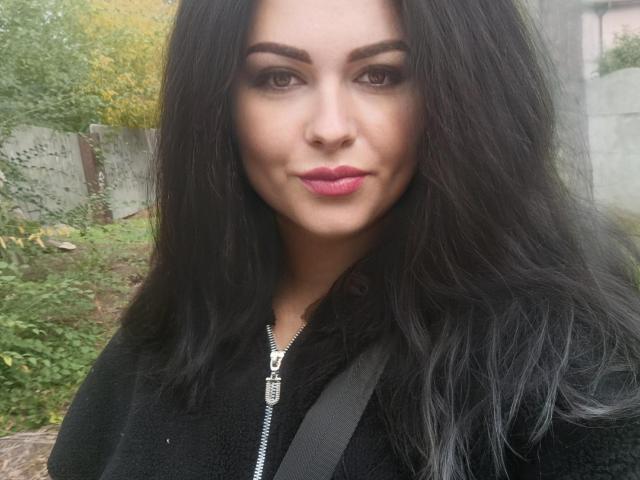 FortuneHelena heeft donker, golvend haar, volle, roze lippen en een lichte glimlach. Ze draagt een zwarte jas met rits en een band over haar borst. Haar bruine ogen zijn gefocust..