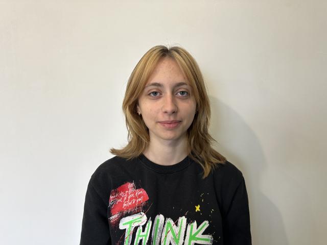 TracyLeex has blonde hair, blue eyes, and freckles, wearing a black "Think" shirt with green and red graphic, standing against a white wall.