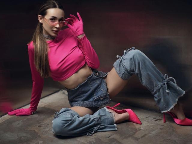 AlexaWilds with pink top, pink gloves, and pink heels, sitting with legs spread, wearing ripped denim shorts, and showing her toned ass. Her long brown hair is down.