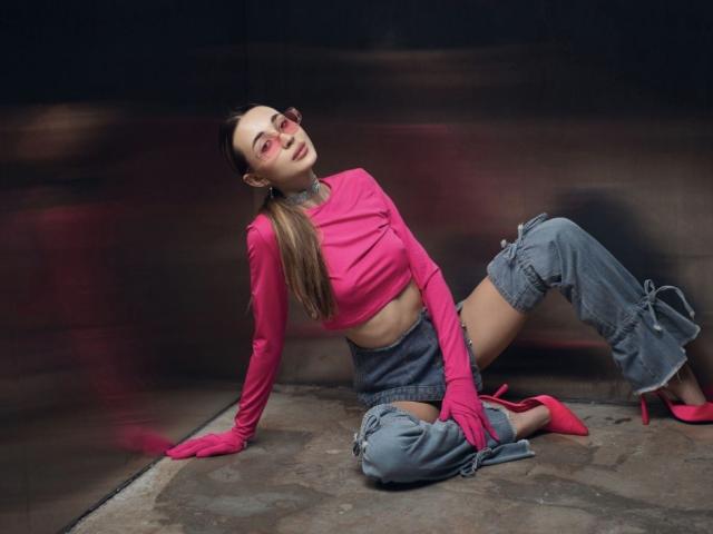 AlexaWilds with pink long sleeves, pink high heels, and blue jeans, sitting on a concrete floor, leaning back, showing her ass, with a blurred background.