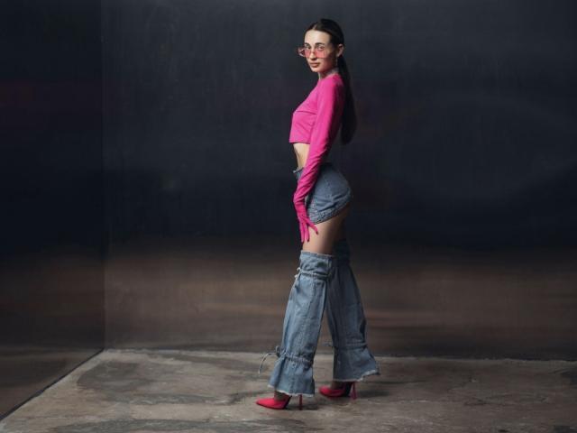 AlexaWilds with long dark hair, glasses, pink top, denim shorts, and blue leg warmers, standing sideways, showing her ass, in red heels, against a dark background.