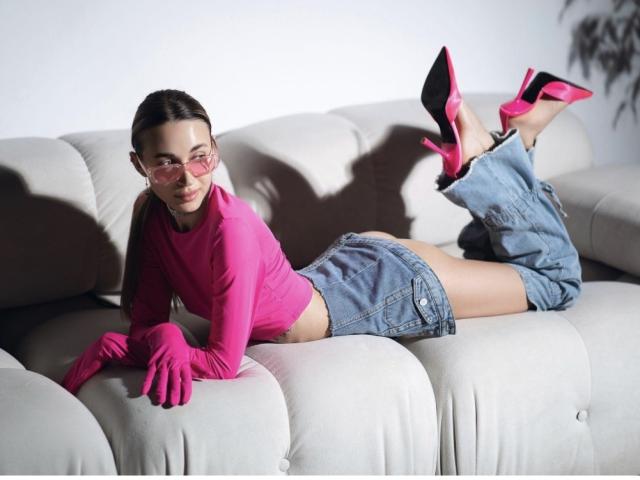 AlexaWilds laying on a white couch, wearing pink glasses, pink long-sleeve top, pink gloves, denim shorts, and pink high heels, with her ass and legs raised in the air.